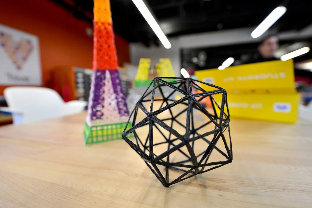 A black geometric 3D model made of intersecting struts sits on a light wood table in a bright office or studio space. A colorful eiffel tower model and a yellow box are blurred in the background.